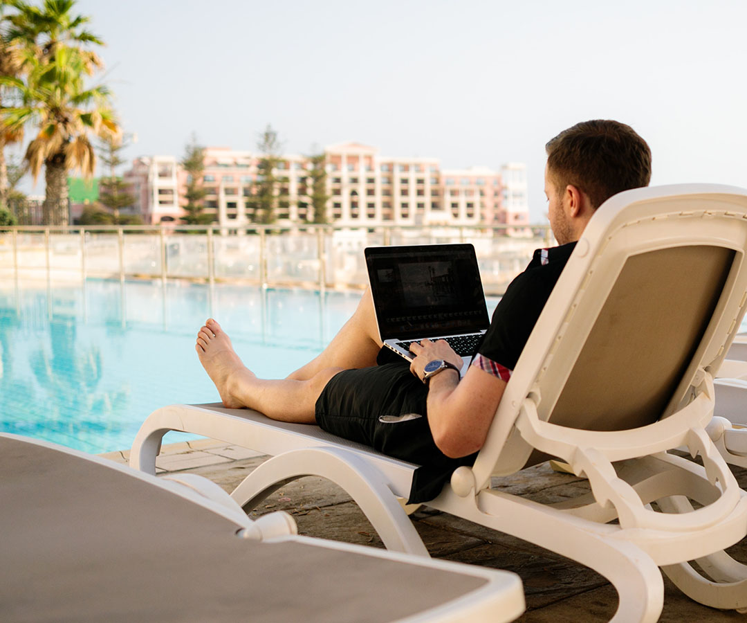 Man on chaise lounge poolside working on laptop